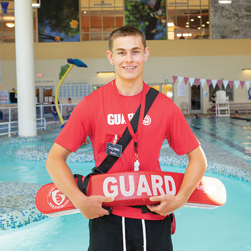 Smiling lifeguard in red shirt stands in front of the pool at the Kroc Center.