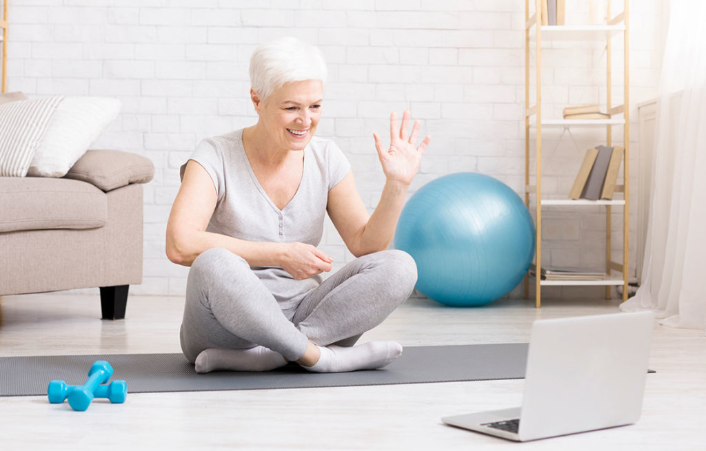 Senior woman sits on a yoga mat with her laptop out, ready to participate in a virtual fitness class.
