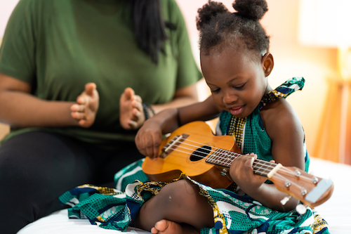 Young girl strums a guitar with her mom sitting beside her.