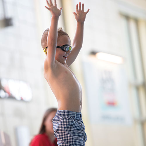 Young boy wearing goggles raises his arms in excitement as he prepares to jump into the pool.