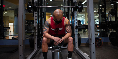 Man in red tank top sits on a bench in front of a weight rack preparing to bench press.